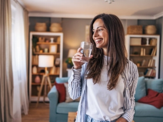 Junge Frau steht im Wohnzimmer und hat Wasserglas in der Hand